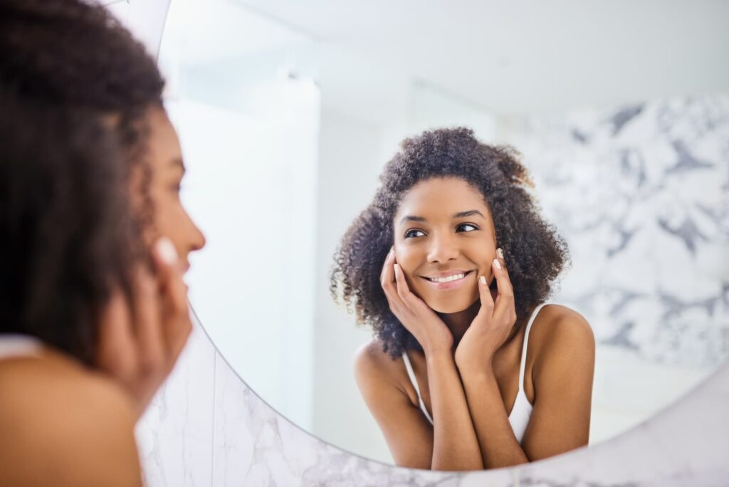 A young woman smiles at herself in the mirror, happy with her soft skin after a series of chemical peels near Macomb.